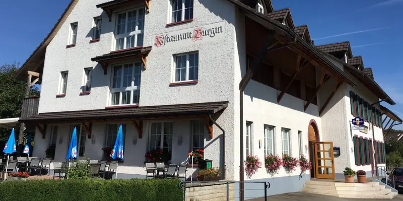 Das Restaurant Bürgin in Wittinsburg von aussen: weisses Haus mit dunklem Holzwerk, Blumenkästen und blauen Sonnenschirmen vor der Terrasse