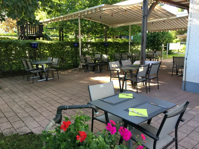 Gartenterrasse unter Bäumen mit gedeckten Tischen und Stühlen, im Vordergrund rote und pinkfarbene Geranien