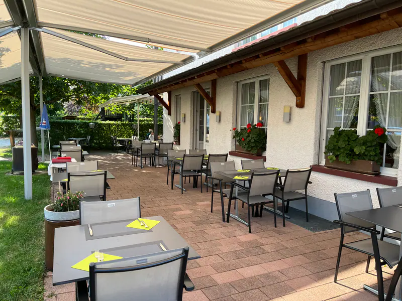 Überdachte Gartenterrasse mit Markise, Tischen und Stühlen, hellgrünen Sets auf den Tischen und Blumenkästen an der Hausfassade