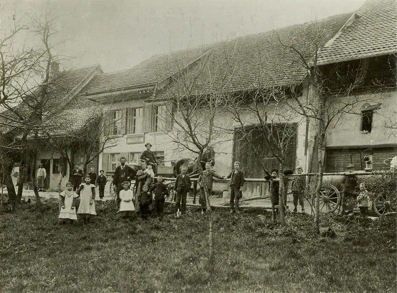 Historische Aufnahme aus der Zeit um 1900: das Bauernhaus, davor Kinder und Erwachsene mit einem Wagen