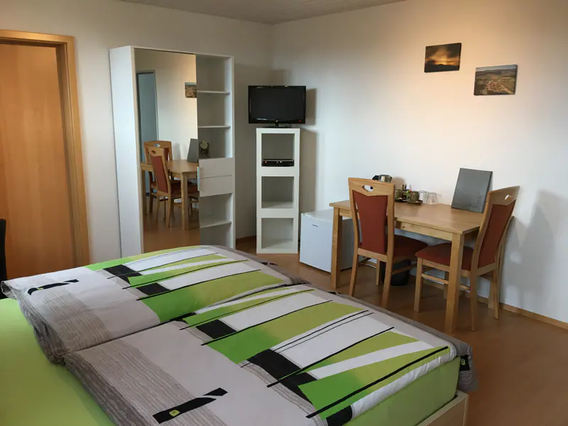 Hell eingerichtetes Hotelzimmer mit Doppelbett, Esstisch mit Stühlen und Fernseher